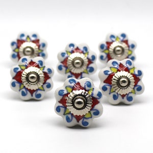 May include: Six white ceramic cabinet knobs with a floral design. The knobs feature red, yellow, and blue accents and a silver metal center.