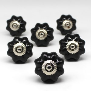 Könnte beinhalten: Sechs schwarze Keramik-Schrankknöpfe mit silbernem Blumendesign. Jeder Knopf hat eine erhabene silberne Mitte mit einer kleinen silbernen Kugel in der Mitte.