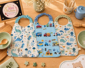 May include: Three fabric bibs with elasticized necklines. The left bib features a white background with blue dinosaurs and trees. The center bib has a light blue background with colorful vehicles. The right bib displays a white background with blue sea creatures.