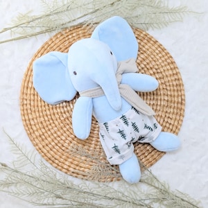 May include: A light blue plush elephant toy with large ears, wearing a gray scarf and white shorts with a green tree pattern. The elephant is lying on a woven, circular mat, with decorative sprigs around the edges.