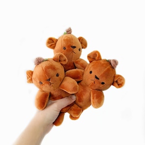 May include: Three plush brown teddy bears with small ears and embroidered facial features. Each bear has a small, light brown hat with a green stem. The bears are held in a hand against a white background.