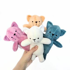 May include: Four plush cat toys in different colors: pink, white, light blue, and orange. Each cat has black eyes and a black nose.