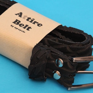 May include: A black recycled tire belt with a silver buckle and two silver rivets. The belt is rolled up and wrapped in brown paper with the text "Attire Belt by up-cycle" printed on it.