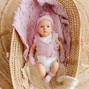 May include: A baby wearing a pink knitted hat, vest, and booties, lying in a wicker basket lined with a white knitted blanket.