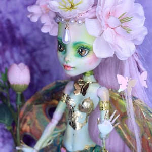 May include: A custom doll with a green complexion, adorned with a floral headdress and gold accents. The doll features a detailed bodice and a skirt with jewel-like embellishments. The doll holds a flower and has butterfly accents.