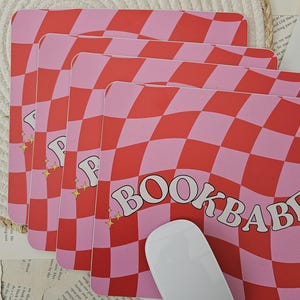 Puede incluir: Una alfombrilla de ratón a cuadros rosa y roja con el texto "BOOKBABY" en una fuente retro. La alfombrilla de ratón es ondulada y tiene un ratón de ordenador blanco en la parte superior.