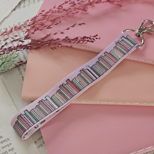 Può includere: Un cordino rosa chiaro con chiusura argentata, con un motivo ripetuto di libri colorati. Il cordino è appoggiato su quaderni rosa e pagine di libri, con fiori rosa secchi sullo sfondo.