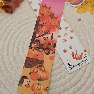 Op de afbeelding: Twee herfst-thema bladwijzers met illustraties van herfstbladeren en pompoenen. Eén bladwijzer toont een tractor en pompoenen, de andere heeft bladeren en de tekst "BookdragonMarit". Decoratieve herfstbladeren en een pompoensticker zijn verspreid.
