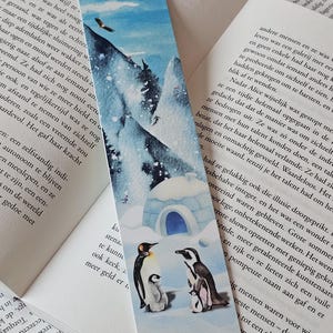 May include: A watercolour bookmark depicting a snowy mountain landscape with an igloo and penguins. The bookmark has a blue sky with a bird and is set against a book's pages.