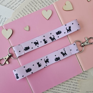 Peut inclure: Deux porte-clés en tissu rose avec des silhouettes de chats noirs et de petits cœurs blancs. Les porte-clés sont attachés à des anneaux de clés argentés. Les porte-clés sont posés sur un livre avec du texte.