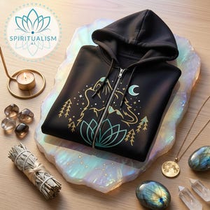 May include: Black zip-up hoodie with a wolf and mountain graphic in gold and teal. The hoodie features a hood and a silver zipper. The image also includes crystals, a candle, and a necklace with a moon pendant, suggesting a spiritual theme.