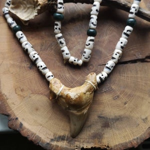 May include: A handcrafted necklace featuring a large, light brown shark tooth pendant. The necklace is strung with white beads, each adorned with a skull design, and interspersed with dark green beads. The necklace rests on a wooden surface.