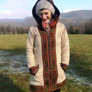 Puede incluir: Una chaqueta con capucha de color crema con cierre de cremallera y detalles bordados coloridos. La chaqueta tiene una cremallera marrón y un patrón bordado marrón y verde en la parte delantera.