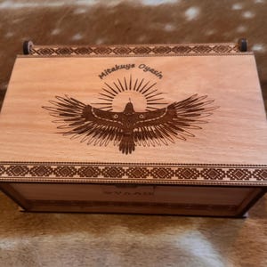 May include: Wooden box with a hinged lid. The lid features a laser-engraved design of a bird with outstretched wings and the text "Mitakuye Oyasin".