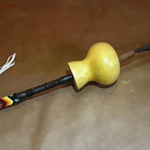 Può includere: Un sonaglio fatto a mano con un corpo di zucca e un manico di perline. Il manico è nero con un motivo geometrico colorato. Un cinturino in pelle bianca è attaccato al manico. Il sonaglio ha anche una coda marrone scuro.