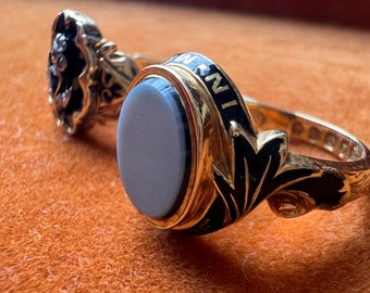 Puede incluir: Dos anillos de oro con esmalte negro y una piedra blanca. El anillo de la izquierda tiene un diseño floral y el anillo de la derecha tiene un diseño más ornamentado con una piedra blanca.