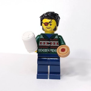 May include: A Lego minifigure with a black hairstyle and an eye patch, holding a white coffee cup and a donut. The figure wears a green sweater with a red and white pattern, a blue shirt, and blue pants.