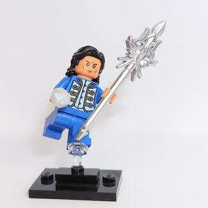 May include: A blue Lego minifigure with black hair, a blue and gold jacket, and a silver staff with a star-shaped top. The minifigure is holding a clear gem in one hand and the staff in the other. The minifigure is standing on a black Lego base.