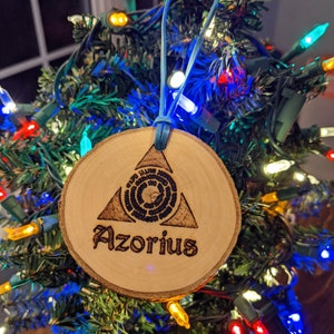 May include: A round wooden Christmas ornament with the word "Azorius" burned into the wood. The ornament features a triangle and circular design. A blue ribbon is attached for hanging. The ornament is displayed on a Christmas tree with colorful lights.