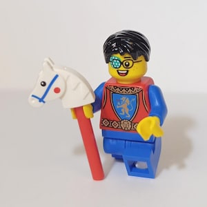 May include: A Lego minifigure of a knight with a red and gold chest plate, blue pants, and a black eyepatch. The knight is holding a white toy horse on a red stick.