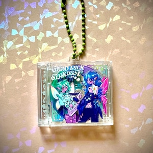 Puede incluir: Un llavero acrílico transparente con dos personajes de estilo anime con alas de hada. El llavero tiene una cadena de cuentas verdes y el texto "GOOD LUCK STARDUST". Los personajes son en tonos azules, verdes y rosas.
