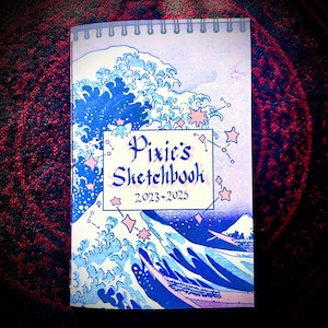 May include: A spiral-bound sketchbook with a cover featuring a blue and white wave design, stars, and the text "Pixie's Sketchbook 2023-2025". The notebook has a light purple spiral binding.