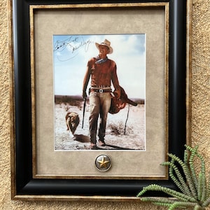Peut inclure: Une photographie en noir et blanc encadrée d'un homme portant un chapeau de cowboy et une veste en cuir marron, tenant un fusil et une selle. L'homme marche avec un chien à ses côtés. La photographie est signée "Clint Eastwood".