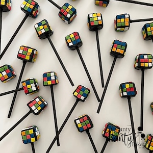 May include: A collection of colourful cake pops shaped like Rubik's Cubes on black sticks. The cake pops are decorated with red, blue, green, yellow, orange, and white squares.