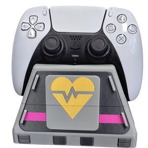 May include: A white and black PlayStation 5 controller rests on a gray charging stand. The stand features a gold heart design with a pink stripe. The controller has black joysticks and button icons.