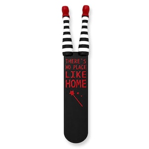 May include: A whimsical bookmark featuring the legs of a witch, with red shoes and black and white striped stockings. The black bookmark has the text "THERE'S NO PLACE LIKE HOME" in red, along with a star and wand design.