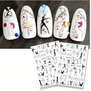 May include: White nail art with ballet dancer designs and rhinestones. The nails feature the word "Graceful" and ballet poses in black, pink, and yellow. Two sheets of nail art stickers with ballet dancer designs are also visible.