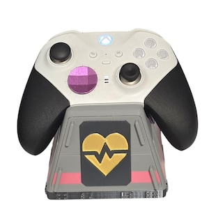 May include: A white and black Xbox controller resting on a gray charging stand. The stand features a black panel with a gold heart design and pink accents. The controller has black grips and a purple center button.