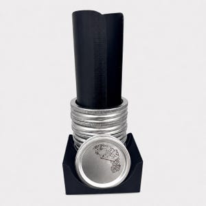 May include: A black plastic stand holding a stack of silver metal mason jar lids. The top lid features a black line art illustration of various fruits and vegetables. A tall, curved black plastic sheet stands vertically inside the lid stack.