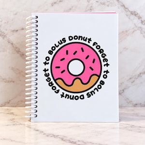 May include: A white spiral-bound notebook featuring a central graphic of a pink frosted donut with brown dough and black sprinkles. The text "Donut forget to bolus" is printed in a circular black font around the donut illustration on the cover.