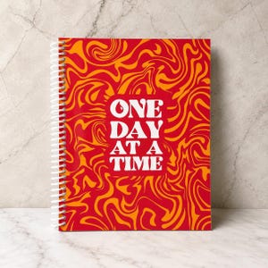 May include: A spiral-bound notebook featuring a vibrant red and orange marbled pattern across the cover. Centered on the front is a white rectangular block containing the bold white text that reads ONE DAY AT A TIME.