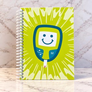 May include: A spiral-bound notebook featuring a lime green cover with a central graphic of a smiling blood glucose monitor. The monitor is lime green and dark blue with a white screen displaying a smiley face, arrow buttons, and the letter M.