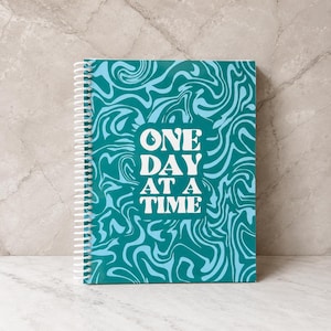 May include: A spiral-bound notebook featuring a teal and light blue marbled swirl pattern on the cover. Centered on the front is a white rectangular graphic containing the text ONE DAY AT A TIME in a bold, stylized white serif font.