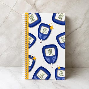 May include: A spiral-bound notebook with a white cover featuring a repeating pattern of blue blood glucose monitors. Each monitor displays the text "YOU GOT THIS!" on its screen, accompanied by small yellow star icons scattered throughout the design.