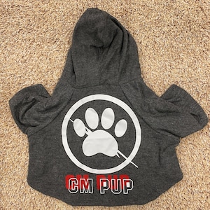 May include: Gray hooded dog sweater with a white paw print inside a circle with a diagonal line through it. The text "CM PUP" is printed in red and white below the paw print.