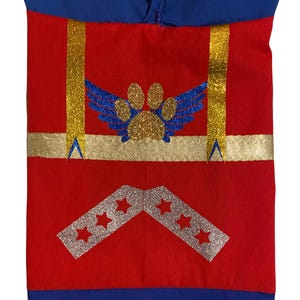 May include: Red fabric dog costume with gold and silver glitter accents. The costume features a paw print with wings and a chevron design with stars.