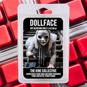 May include: A clear plastic clamshell package containing a soy blend wax melt. The label features the text DOLLFACE, SOY BLEND WAX MELT 2.4 OZ (68 g), THE KINE COLLECTIVE, and AGGRESSIVELY GOOD-SMELLING HOME FRAGRANCE FROM LANCASTER, PENNSYLVANIA.