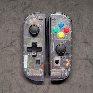 Puede incluir: Un par de mandos Nintendo Switch Joy-Con transparentes grises con botones de colores. Los mandos están separados y muestran el circuito interno.