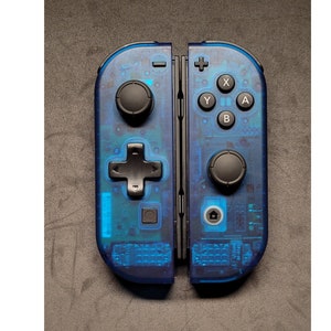 Puede incluir: Un par de mandos Nintendo Switch Joy-Con transparentes azules con botones negros. Los mandos están separados y muestran el circuito interno.