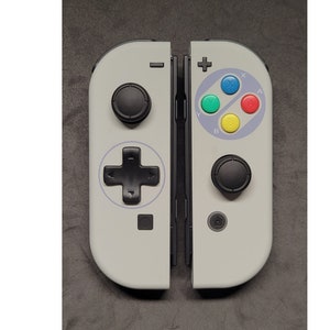 Puede incluir: Un mando blanco de Nintendo Switch con un diseño Super Nintendo gris y negro. El mando tiene un pad direccional negro, cuatro botones de colores y un botón de inicio negro.