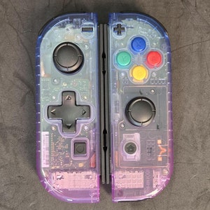 Puede incluir: Un par de mandos Nintendo Switch Joy-Con transparentes de color morado con botones negros. Los mandos son translúcidos y muestran los componentes internos.