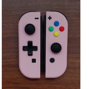 Könnte beinhalten: Zwei rosa Joy-Con-Controller für eine Spielkonsole mit schwarzen Knöpfen und Richtungspads. Der rechte Controller hat farbige Knöpfe in Rot, Grün, Blau und Gelb. Die Controller liegen auf einer braunen Holzoberfläche.