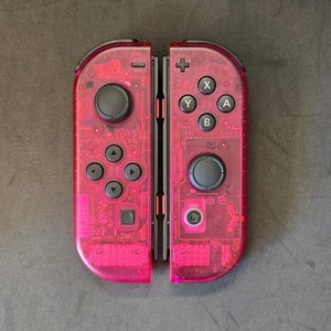 Puede incluir: Un par de mandos Joy-Con Nintendo Switch de color rosa. Los mandos son transparentes y muestran el circuito interno. Los botones son negros y están etiquetados con las letras X, Y, A y B.
