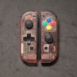 Puede incluir: Un par de mandos de juego rosas transparentes, que muestran los componentes internos. Cada mando tiene joysticks y botones negros, con botones de colores en un lado. Diseñados para jugar.