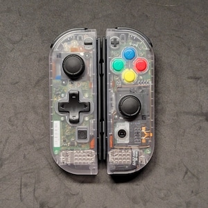May include: A clear, transparent Nintendo Switch controller with black buttons and a visible circuit board. The controller is in two parts, with the left and right sides separated.