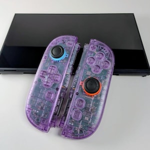 Puede incluir: Dos mandos de juego morados transparentes con componentes internos visibles se colocan delante de una pantalla negra. Los mandos tienen detalles en negro, azul y naranja. Los mandos están diseñados para jugar.